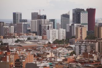 Vista aérea panorâmica de Barcelona, com edifícios residenciais e arranha-céus formando a paisagem urbana.