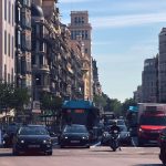 Rua movimentada de Barcelona com ônibus, carros e motos diante de edifícios urbanos.