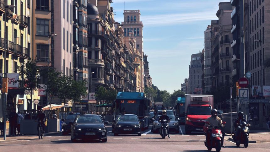 Rua movimentada de Barcelona com ônibus, carros e motos diante de edifícios urbanos.