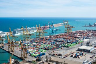 Vista aérea do Porto de Barcelona, com guindastes, contêineres e navios em uma área de operações portuárias.