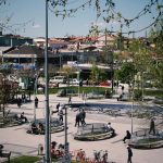 Pessoas caminham e descansam em um parque urbano arborizado, cercado por edifícios.