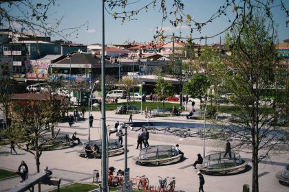 Pessoas caminham e descansam em um parque urbano arborizado, cercado por edifícios.